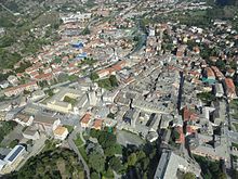 Altra veduta aerea di Susa. Sono ben visibili la Cattedrale di San Giusto e il suo campanile affiancata dal vecchio seminario a sinistra e da Porta Savoia sulla destra, i quali si affacciano su Piazza Savoia, (dove sono anche collocati gli scavi archeologici), e la Pieve battesimale di Santa Maria Maggiore che si affaccia sul cortile dell’oratorio della parrocchia. Accanto al ponte sulla Dora Riparia si nota la Chiesa della Madonna del ponte, sede del Museo diocesano d’arte sacra. Si scorge in lontananza (sulla sinistra) anche il campanile color giallo della Chiesa di Sant’Evasio e dietro di questa l’edificio che ospitava la sede segusina del Cotonificio Vallesusa. In basso a destra si può notare il tetto del Castello della Contessa Adelaide