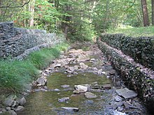 A stream flows over smooth rocks between walls of rock held in place with wire. Grass and trees grow on the banks at the top of the walls.