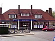 A brown-bricked building with a red-tiled roof and a rectangular, dark blue sign reading "WATFORD STATION" in white letters all under a white sky