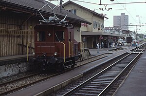 Red shunting electric locomotive in foreground of three-story station building with flat platform canopy