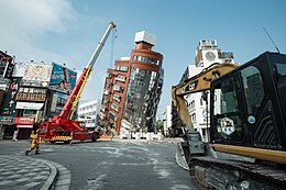 A large building at center leans forward. Heavy machinery is visible in the midground and foreground. A man walks at left.