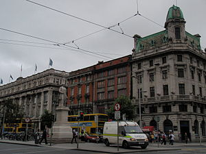 A busy street with large buildings, many cars and tram power cables overhead.