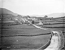 Photographie en noir et blanc d'un paysage de campagne, à gauche et à droite des champs de culture séparés par des murets de pierre, entre les deux, une route où circule une charrette, au fond des habitations
