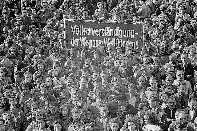 "Schwarz-Weiß-Fotografie zeigt Menschenmenge und ein Plakat mit der Aufschrift: Völkerverständigung – Der Weg zum Weltfriedenǃ"