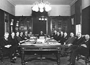 A black-and-white photo of fourteen people sitting around the table in an official setting