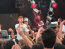 Tom Scott, onstage at a music festival, speaks into a microphone. Other members of his band are visible around him.