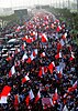 From above, an endless sea of people holding red and white flags are marching in the middle of a city highway surrounded by palm trees