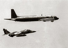 Black-and-white photography of jet aircraft, carrying external fuel tank under fuselage, accompanying behind a four-turboprop-driven aircraft.