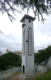 Fotografía de una gran torre blanca coronada por un techo piramidal.  En su fachada izquierda hay una puerta de entrada en la parte inferior y un reloj en la parte superior.  En la parte superior del techo hay una veleta.