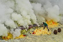 En mann i enkle støvler arbeider svovel på bakken med en pæl på kanten av fumaroles.