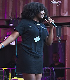 Noname onstage smiling, holding a microphone