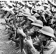 Un grand nombre de soldats asiatiques bien équipés assis en rangées sur le sol, portant des casques coloniaux et tenant des fusils d'assaut.
