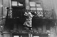 "A woman hangs from a balcony, preparing to drop to the street and the waiting SS."