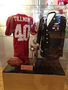 Uma camisa e uma bola de futebol americano, um troféu Pat Tillman e uma jaqueta militar exibidos sob uma vitrine de vidro.