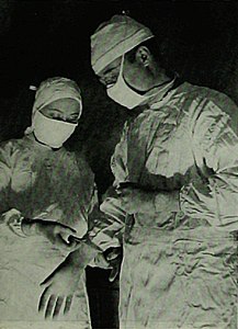 "Doctor Freeman is the neurologist and analyst, Doctor Watts the surgeon. Here the latter is being helped on with his gloves before operating."