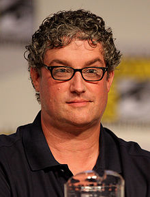 A man with glasses and a black shirt sits in front of a microphone.