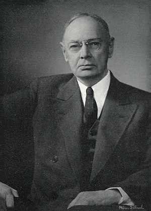 Formal black and white portrait of an older man, wearing glasses, in a suit