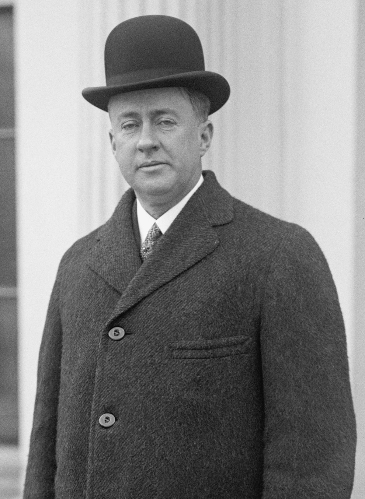 Black and white image of a clean shaven man with drooping eyelids， aged about forty. He is wearing a black bowler hat， white shirt， tie and dark overcoat.