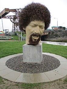 Sculpted head of a man with goatee and curly hair presented on a block. The pavement around it is circular. In the background, there is a neatly manicured lawn, and construction crane. There is a walkway and stream on right corner.
