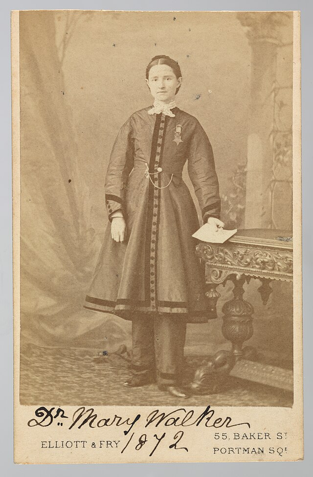 In this monochrome brown and white image, a white woman poses at center. Her complexion is white, with her hair braided and pinned close to her head. She wears a buttoned overcoat that ends at mid-calf with detailed banding on the hem and at wrists, and matching long pants beneath. She stands at center, on a brocade carpet, in front of a painted backdrop, her left hand rests on a carved wooden table, holding a document, her right hand rests in front of her coat. The printed image is mounted on a white card, which is annotated at the bottom "Dr. Mary Walker 1872" in manuscript ink, above and between the printed name and address of the photograph studio [55 Baker Street, Portman Square]