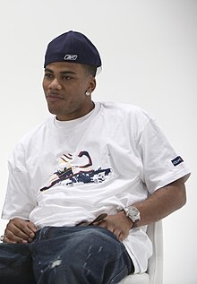 An African man sitting on a chair and looking to the right of the image. He wears white T-shirt and navy blue jeans and cap, topped with a watch on his left hand.