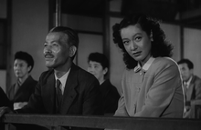 Een theater waar een Noh-toneelstuk wordt opgevoerd: Prof. Somiya draagt ​​een pak en stropdas en Noriko draagt ​​een eenvoudige, westerse jurk met kraag;  Shukichi kijkt recht voor zich uit naar het linkerframe van de foto, glimlachend, en Noriko, niet glimlachend, kijkt naar het rechterframe van de foto naar een onzichtbare persoon.