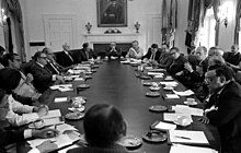 Tyve mennesker mødes i et mødelokale omkring et ovalt bord.  En mand, i midten af ​​bordet i højre side, henvender sig til de andre.  Alle er iført jakkesæt eller lignende påklædning.