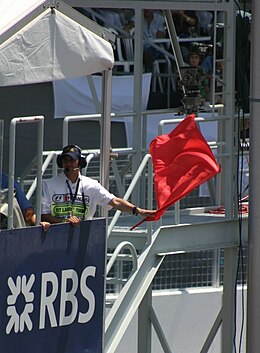 A man in a white overall, standing behind a blue sponsorship advertisement for the Royal Bank of Scotland, with the letters "RBS" in white. He is waving a red flag.