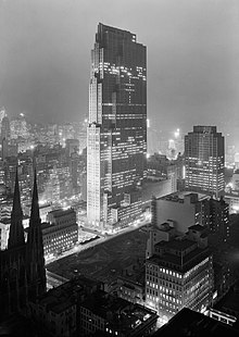 A monochrome image of a building being constructed, with lit office rooms especially prominent