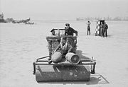 Two men drive a jeep, pulling a ground roller, on which a third man sits. Other men watch in the background. Parked aircraft can be seen in the distance.