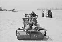 Dos hombres conducen un jeep, tirando de un rodillo de tierra, sobre el que se sienta un tercer hombre.  Otros hombres miran al fondo.  Los aviones estacionados se pueden ver en la distancia.