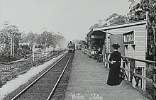 A lady standing on a station platform looks at the camera while a train pulls in from in the distance