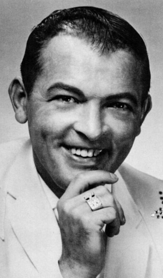 A dark-haired man smiling wearing a white jacket, smiling broadly and holding one hand to his chin