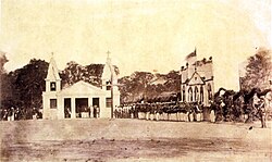 Photograph showing lines of soldiers assembled in a square facing a large wooden cross in the center and a low building or pavilion with 2 flanking spires hung with bells in the background