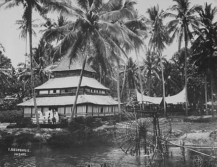 Pajakoemboeh scene with water wheel, children swimming, a mosque and a Minangkabau town hall in the background