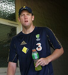 A man is peering in front of him, holding a drinks bottle with a yellow liquid in.  He is wearing a dark blue t-shirt with gold piping, and four logos on it.  He is also wearing a baseball cap of the same colours.