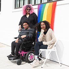 Three Black and disabled people in front of a pride flag. Disabled BIPOC in front of pride flag.jpg
