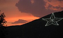 A five-pointed star made up of white lights on a mountain during an orange sunset, with a tree silhouette in the foreground