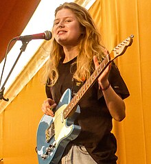 Onstage, a young woman with long bleached hair and a nose ring holds an electric guitar while dressed casually in a t-shirt and jeans