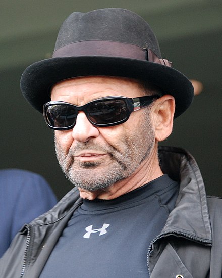 A head shot of Pesci, unshaven, wearing dark sunglasses, hat, shirt, and leather jacket