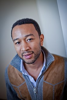 John Legend de frente, olhando para a câmera com um leve sorriso, vestindo uma jaqueta de camurça.