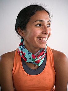 Head and shoulders photo of Jordan Marie Brings Three White Horses Daniel, a young Lakota woman, smiling wearing an orange sleeveless shirt and a teal and red scarf