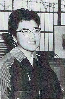 Black and white photo of a woman with short hair. She is wearing glasses and smiling. She is looking at someone outside of the photograph to the right.