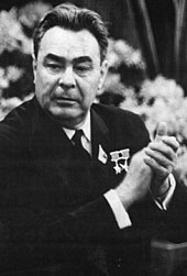 A black and white photograph of a man in a black suit bearing medals on the left breast, applauding as he looks to his right.