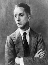 A black-and-white portrait photo of a young Norman Rockwell with his arms crossed in a light suit coat with a dark tie and white shirt