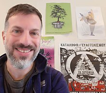 Image of a man with brown hair and grey beard smiling in front of a wall with many colourful activism posters