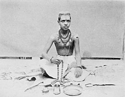 This image shows a young Brahmin Priest sitting on floor with folded feet, "tilak" on his forehead, [[Rudraksh]] mala around his neck, [[Mala]] in his hand and other Pooja ritual necessities laying around him.