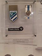 תג כתף שהיה נענד על ידי סגל משמר האזרחי ומתנדבים על מדים ועיטור ההתנדבות המוצגים בבית מורשת משטרת ישראל