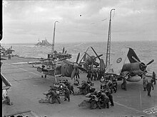 Black and white photograph of several groups of men bent over bombs on the flight deck of an aircraft carrier at sea. Several monoplane aircraft are parked on the flight deck, and another warship is visible on the sea near the carrier.