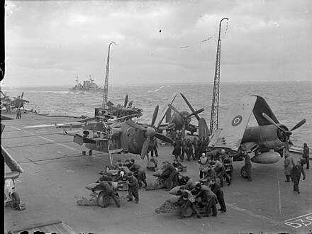 Barracudas being "bombed-up", during Operation Goodwood in August 1944. The heavy cruiser Berwick is in the background.